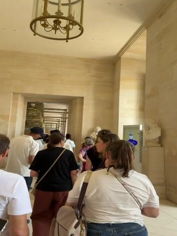 A person's hand holds a smartphone in the foreground, the screen illuminated and pointed toward a hallway. The hallway is lined with large stone blocks and features a grand chandelier on the ceiling. Several people are visible, standing and walking in a line within the hallway, appearing to be in a museum or a similar public building. One woman in the foreground faces away from the camera, wearing a white shirt and jeans, with her back to the viewer. Another woman in a black shirt and reddish-brown pants stands further down the line, also facing away. Towards the middle of the hallway, a young woman with brown hair pulled back in a ponytail is looking to her right, engaged in conversation. In the background, through an archway, more people and displays are visible. To the right of the hallway, a tall window with multiple panes is visible, letting in natural light. Next to the window, a white pedestal holds a bust of a person. Further down the hallway, two large white marble statues are displayed on a raised platform. The statue on the left depicts a seated female figure holding a horn-like object, while the statue on the right shows a seated female figure wearing a helmet and holding a bouquet of flowers. The overall environment suggests an indoor public space, likely a museum or historical building, with an emphasis on classical architecture and art.