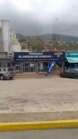The camera is mounted on a vehicle traveling on a road past several buildings and a parking lot. 

In the first shot, a green car is in a parking lot alongside other cars and a tan SUV. A restaurant with signage that includes “PAN” and “El Meson de” is visible.

The camera moves past a building with a sign that includes the words “ALCALDIA BOLIVARIANA DEL MUNICIPIO VARGAS” and “SUPER MERCADO.” A tan SUV is parked nearby. 

The camera passes a building with a blue exterior, gray accent columns, and several windows. 

Finally, the camera passes an outdoor area with red columns and a white roof. People are sitting on benches and walking through the area. A police officer in uniform stands nearby.