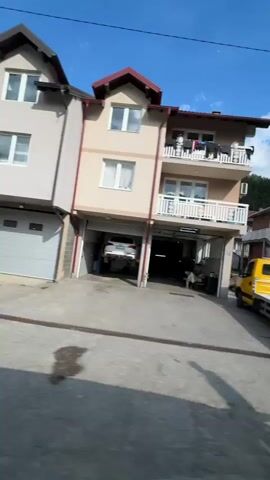 The video shows a quick pass through a residential area. Starting next to what appears to be an auto repair shop, with a car lifted on a hydraulic lift, the camera moves past a series of residential houses on a gravel road. The houses are a mix of brick and plaster, with balconies and varying architectural styles. Laundry hangs on some balconies. There are several parked cars, including a blue van, a red car, and a white van. The sky is blue with scattered clouds, and there are trees visible in the background. The movement suggests the video was taken from a moving vehicle.