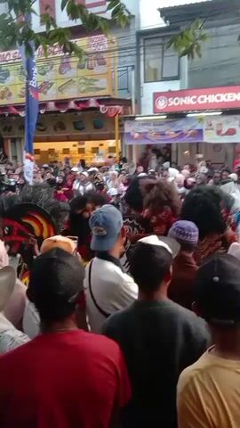 A large crowd of people are gathered on a street, watching a performance. In the foreground, several people are holding up their phones to record the event. The performers appear to be wearing elaborate costumes with large, furry headdresses and face masks. One performer has a long, flowing tail that is visible. Behind the performers, there are buildings with signs advertising various businesses, including "PROMO GUFF", "Bank BRI", and "SONIC CHICKEN". There is also a stage with speakers set up, suggesting a musical performance is taking place. The overall atmosphere appears festive and crowded.