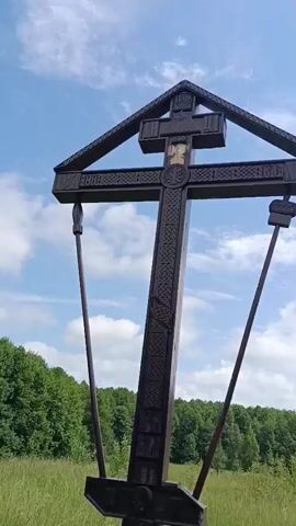 A wooden cross with intricate carvings stands in a grassy field under a blue sky with white clouds. The cross is tall and has an ornate design. There are two thin metal poles attached to the sides of the cross, extending outwards and downwards. A black plaque with text is affixed to the base of the cross. In the background, a line of green trees stretches across the horizon. The camera pans slightly, showing different angles of the cross and its surroundings.