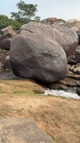 The camera captures a static shot of a rocky landscape with a small stream flowing between and under large boulders. The boulders are predominantly gray and brown, with visible textures indicating weathering. Green grass and sparse vegetation are visible on the surrounding dirt ground and around the rocks. In the background, trees with green foliage are seen against a bright, slightly overcast sky. The stream appears shallow and moves swiftly across the rocks.
