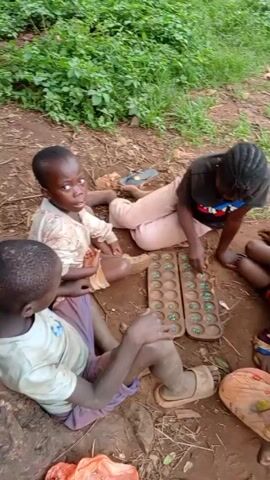 The video shows a group of four children sitting on the ground around a game of Mancala. The game board is wooden with two rows of circular depressions filled with green marbles. The children are of African descent. They are wearing various casual clothes and sandals. The ground is dirt with some patches of grass. In the background, other people can be seen walking around. The children appear to be engaged in playing the game, with one child actively moving the marbles. The atmosphere seems relaxed and playful.