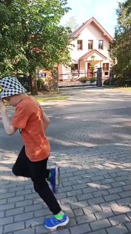 This short video clip captures a young person, likely a child, walking briskly on a brick-paved sidewalk. They are wearing a checkered bucket hat, an orange t-shirt, black pants, and bright blue sneakers with green accents. In their right hand, the child holds a yellow object, possibly a toy or a prop, that they are shaking.

The setting appears to be a suburban or semi-rural area, with a residential house visible in the background. The house is light-colored with a red-tiled roof and visible windows. A fence and gate line the property. Lush green trees with full foliage flank the street and the house, suggesting it is daytime during a warmer season. The sun is casting shadows on the ground, indicating it's likely mid-morning or afternoon.

Cars are seen passing by on the asphalt road adjacent to the sidewalk. In one frame, a black SUV is visible, and in another, a white van. The child's movement suggests they are either on their way somewhere or engaged in a playful activity, possibly imitating or playing with the yellow object they are holding. Their body language, with a bent-over posture and energetic stride, conveys a sense of purpose or enjoyment. The overall mood of the video is bright and active, characteristic of an outdoor daytime scene. No discernible text is visible in the clip. The location context of Kamionka, Poland, places this scene within that specific geographical area.