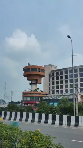 The video shows a tall, cylindrical building with a wider top section and a narrower middle section. The bottom of the building has storefronts, one of which displays a "KFC" sign. The building is situated next to a larger, multi-story building with numerous windows. In the foreground, a black and white striped barrier runs along a road, and a blue truck with a cargo container is driving past. The sky is overcast.