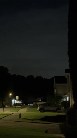 The video shows a nighttime suburban scene. The sky is dark with some faint clouds. In the foreground, there is a grassy lawn with a sidewalk. Further back, there is a street with a few parked cars, houses, and trees. A street light illuminates a portion of the sidewalk. In the distance, there is a faint light flash, possibly lightning. The overall ambiance is quiet and still.
