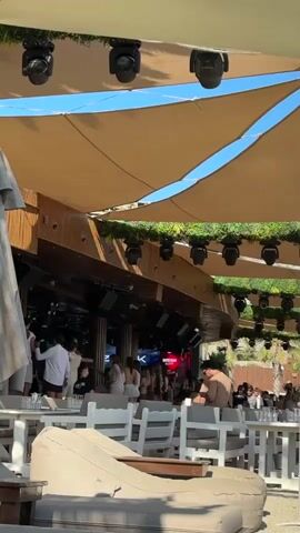 A group of people are gathered under a large tan canopy supported by structures. The canopy is adorned with numerous spotlights and has greenery along its edges. Below the canopy, a building with wooden paneling is visible, featuring more lights and what appears to be a bar area. Several individuals are standing and interacting in this area, some facing a screen that displays colorful graphics. In the foreground, tables with white chairs and a large, light-colored beanbag chair are arranged, suggesting an outdoor seating or dining space. The overall scene depicts a lively outdoor venue, possibly a beach club or an entertainment establishment.