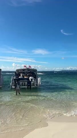 This media captures a vibrant scene at a beach, likely in Phru Nai, Thailand, under a bright, sunny sky with scattered white clouds. The turquoise waters of the ocean gently lap against a pristine white sandy beach in the foreground.

In the middle ground, a white boat with a canopy is anchored close to the shore. Several people are visible on and around the boat, with some appearing to be preparing to disembark or having just arrived. The boat is equipped with outboard motors at the stern. Two Thai flags are flying from the boat, indicating its origin or affiliation.

Further out in the water, a line of jet skis is visible, suggesting a recreational area or water sports rental facility. Some jet skis are stationary, tethered in a line, while others might be in use or waiting for riders. The water is clear, allowing a glimpse of the seabed in shallower areas. In the distance, a chain of islands or coastal mountains forms the horizon line under the expansive blue sky.

The overall atmosphere is one of leisure and vacation. The clear weather and the presence of boats and jet skis point to a popular tourist destination during daylight hours, possibly mid-morning or early afternoon, given the bright sunlight and clear visibility. The scene evokes a sense of tropical relaxation and outdoor fun. No visible text is present in the media.