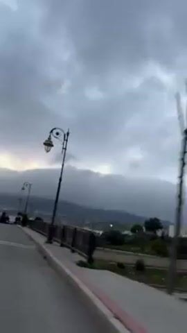 The video shows a cloudy day, with overcast skies. There is a road with street lamps and a black metal guardrail. In the background, there are hazy mountains and buildings in the distance. People walk along the side of the road.
