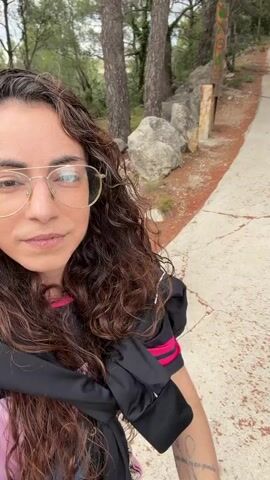 A woman with curly brown hair and glasses takes a selfie in front of a narrow, paved path with trees in the background. Another woman sits to the right of the path, holding a paper bag of food and looking at her phone. A white SPAR shopping bag sits next to her. The environment appears to be a park or wooded area.