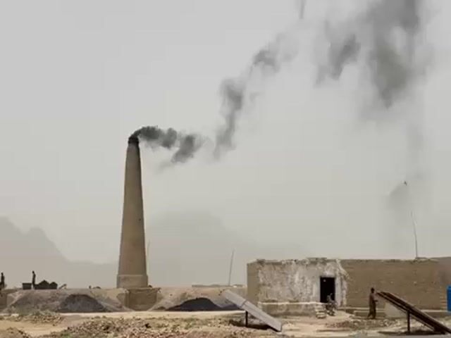 Here's a description of the video:

The video shows a brick factory with a tall chimney billowing thick black smoke into the hazy sky. In the background, rugged mountains are visible. The factory structure appears to be made of brick or mud brick, with a small building next to the chimney. In the foreground, there are piles of what appear to be raw materials or fuel. People can be seen moving around near the base of the chimney. A motorcycle carrying at least two individuals passes in front of the factory. The overall scene suggests a harsh, dusty environment. There appears to be a solar panel in the foreground as well.