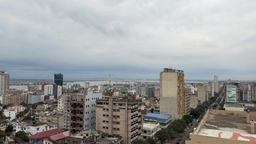 An aerial panoramic view of a city skyline with a bridge in the distance. The sky is overcast with heavy clouds. Buildings are densely packed, varying in height and architectural style. Some buildings appear to be under construction or in disrepair, with exposed concrete and unfinished facades. In the foreground, older, low-rise structures with balconies are visible. In the mid-ground, taller buildings, including apartment blocks and office buildings, dominate the cityscape. A wide street lined with trees runs parallel to a row of buildings. In the background, a large suspension bridge spans a body of water, with ships docked at a port. The overall impression is of a developing urban environment.
