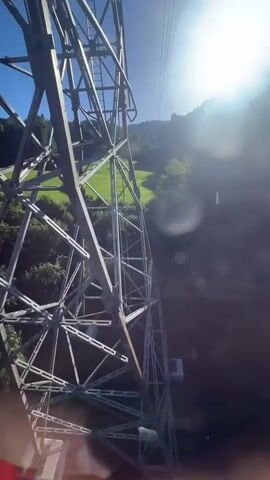 This is a first-person perspective video shot from a ski lift going up a mountain. The ski lift structure is visible in the foreground, made of metal beams. Below the ski lift, a lush green grassy slope is visible, with some trees and a wooden building. The sun is shining brightly, casting a glare on the lens and creating lens flare. The sky is clear and blue. The person filming is holding onto the metal bar of the ski lift.