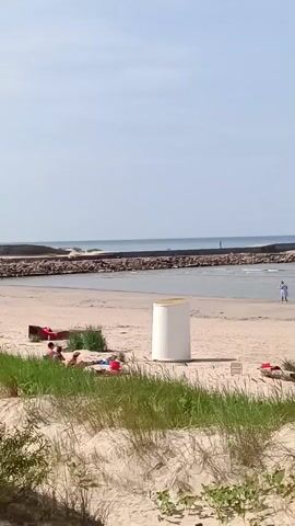 A wide shot shows a sandy beach with green grass in the foreground. A white cylindrical object stands in the middle of the beach. Further back, several people are sitting on the sand. The ocean is visible with a breakwater in the distance, featuring two lighthouses.