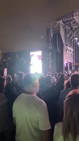 The video shows a concert crowd from behind, facing a brightly lit stage. The stage has a large screen displaying a performer, and band members are visible playing instruments. The text "TIO PEPE FESTIVAL" is emblazoned on the stage rigging. The audience members are diverse, with various hairstyles and clothing. Some are looking towards the stage, while others are turned slightly away. The overall atmosphere appears to be lively and energetic.