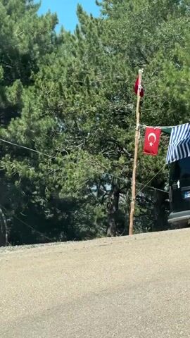 The video shows two brief scenes. The first scene is of a Turkish flag hanging from a wooden pole next to a black van, with pine trees in the background. The second scene is a reflection in a convex mirror, showing a road with a white bus driving away.