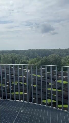 The video begins with a view of a wooded landscape under a partly cloudy sky, seen from an elevated platform with a metal grate floor and a railing made of metal bars and wooden planks. The camera then pans slightly to reveal more of the park-like area below, featuring green grassy fields with winding paths and some structures. The view shifts to a man wearing a light-colored t-shirt and a baseball cap, seen from behind as he looks out at the landscape. In the distance, a red-roofed building with a white steeple is visible amidst the trees. The camera then pans back to show the railing and the landscape beyond, with a circular paved area and a covered structure visible in the middle ground. The video ends with a final view of the landscape.
