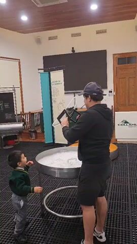 A man in a black hoodie, black shorts, and a baseball cap holds a large bubble-making apparatus. A young child in a green and white school uniform stands to the left, facing a large metal tub filled with what appears to be soapy water. The man is demonstrating how to make bubbles, and the child is watching intently. The room is indoors, with a wooden ceiling, tiled floor, and several display boards and banners in the background. The lighting suggests an indoor event or exhibition.