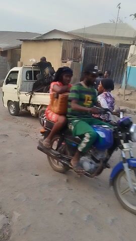 The video captures a street scene in Mpape, Nigeria, featuring an unpaved, dusty road lined with buildings that appear to be houses or shops. Several people are seen riding motorcycles. At the beginning, a motorcycle with two riders passes the camera, followed by another motorcycle carrying three individuals. Later, another motorcycle with two riders also passes by. A small, white utility vehicle, resembling a mini-truck or van, is briefly visible on the side of the road. The atmosphere suggests a typical daytime street with ongoing local traffic and activity under clear skies.