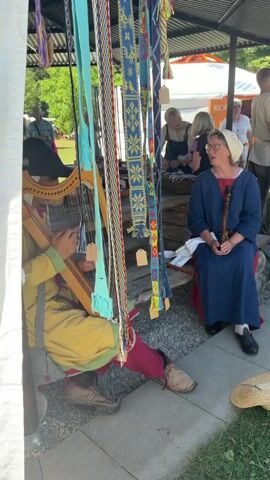 The video opens with a close-up of colorful woven straps hanging down, obscuring a person playing a harp. The person on the harp is wearing a yellow tunic and red pants, with brown leather boots. A woman in a blue dress and a white head covering sits to the right of the harp player, holding a wooden flute. She is looking off-camera to the left. Behind them, more woven straps hang, some with price tags. In the background, there are other people, some browsing at stalls, and tents. The ground appears to be a mix of gravel and grass. The scene suggests an outdoor market or historical reenactment.