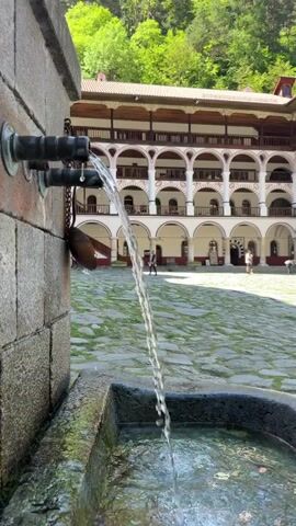 The video shows a courtyard of a multi-story building with arched balconies, filmed from near a stone fountain. Water pours from three metal spouts into a stone basin.  People walk through the courtyard. The building has a white exterior with dark wood trim and is surrounded by trees. The ground is paved with cobblestones.  The sky is bright blue.