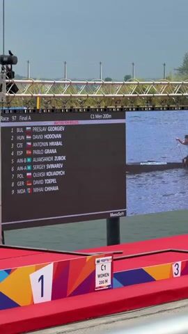 A large screen displays the results of race 97, final A, for C1 men 200m. The top competitor listed is Preslav Georgiev from Bulgaria. The screen also shows a person in a canoe on the water. In the foreground, red platforms with large numbers "1" and "3" are visible, indicating podium positions. The environment appears to be an outdoor water venue, possibly a competition course, with trees and hills in the background. Several other individuals are seen in canoes on the water further in the distance, some of whom are positioned near numbered buoys. A speaker is positioned in the lower right corner of the frame.