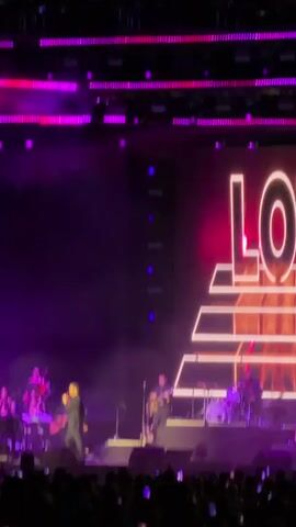 The video captures a live concert scene, featuring a male singer performing on stage with a full band and orchestra. The band consists of guitarists, a drummer, a bassist, and keyboard players. The orchestra, positioned to the side, includes string and wind instruments. The stage is illuminated with purple and pink lighting, creating a vibrant and energetic atmosphere. Behind the performers, a large screen displays a graphic design with bright neon lines. The audience, visible in the foreground, is holding up their phones to record the performance. The lighting is dramatic, with a mix of stage lights and neon graphics that emphasize the lively ambiance of the event.
