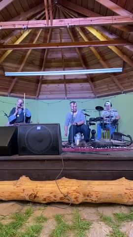 Three individuals are performing music on a raised wooden stage beneath a wooden gazebo with a conical roof. The person on the left, facing forward, is seated and playing a theremin. In the center, another individual is seated and playing a percussion instrument, possibly a frame drum or bongo, with their hands. To the right, a third person is seated and appears to be playing a drum set, with cymbals and drumsticks visible. A large black speaker is positioned to the left of the stage. A string of fairy lights is visible along the back wall of the gazebo. The stage is covered with a red rug. A thick, peeled log runs horizontally in the foreground. The ground in front of the stage is covered with grass.
