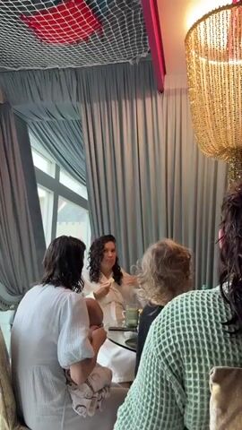 Four women are seated around a table, engaged in conversation. One woman is wearing a white robe and holding a small white object to her mouth with her right hand. Another woman is holding a baby wrapped in patterned cloth. A third woman with curly hair is seated beside them. The fourth woman is seated facing away from the camera, wearing a green sweater. A window is visible behind the group, with light blue curtains covering the majority of the view. A gold chandelier hangs above the table. The ceiling has a decorative net with red and white accents.