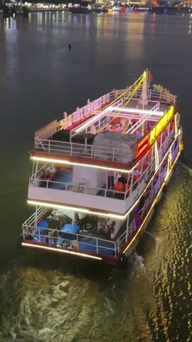 A nighttime view shows a multi-tiered boat sailing on a body of water. The boat, lit with numerous lights, carries passengers on its different levels. Behind the boat, the city skyline is visible, also illuminated. The camera follows the boat as it moves away, leaving a trail of water. There is a buoy visible in the water at one point.