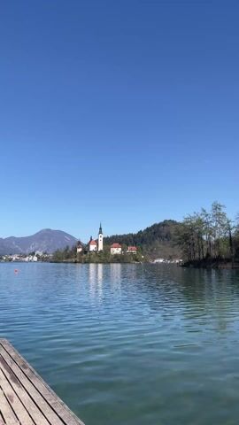 The video shows a scenic view of Lake Bled in Slovenia on a bright, sunny day. The camera is positioned at the edge of a wooden dock, with the calm, turquoise water of the lake filling the foreground. In the distance, a small island with a church and several other buildings is visible. The church has a prominent white tower with a dark spire. A mountain can be seen to the left of the island, and forested hills flank the right side of the frame. The sky is clear and blue, with a faint contrail of an airplane visible high above. The scene is tranquil and peaceful, with the gentle ripples on the water adding to the serene atmosphere.