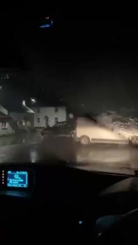 The video is shot from inside a car on a rainy night. Raindrops streak the windshield, obscuring the view somewhat. Outside, there are two vehicles parked on the street, a dark-colored SUV and a silver van. The street is wet and reflects the light from street lamps and the vehicle headlights. In the background, houses are visible, their facades lit by porch lights. The car's dashboard is visible, with the infotainment screen displaying information.  The wipers appear to be running. The scene is dark and wet.