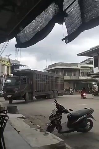 The media file depicts a street scene in Keudegeudong, Indonesia. The primary subject is a large, dark-colored truck with a tall, cage-like cargo bed, positioned in the middle of a paved road. The truck appears to be in motion, with its front wheels turned slightly.

In the foreground, partially obscuring the lower part of the frame, are what appear to be the backs of two chairs, suggesting the perspective is from a seated observer under an overhang. A black scooter is parked on the right side of the foreground.

In the background, several buildings line the street. The architecture suggests a commercial or residential area, with multiple stories and balconies visible. The sky is overcast, indicating a cloudy day, and the lighting suggests it is daytime, likely late morning or early afternoon.

Several other motorcycles and a few people are scattered throughout the background, indicating some level of activity on the street. One person is riding a red scooter in the mid-ground, moving from left to right. The overall atmosphere appears to be that of a typical day in a town or city, with traffic and pedestrian movement.

There is a yellow license plate visible on the front of the truck, but the text is illegible. There is also some text on the building in the background, but it is too blurry to read. The scene is not overtly emotional, presenting a neutral, observational view of the environment.