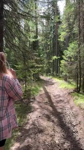 The video shows a person with blonde hair wearing a pink and white plaid jacket and black shorts walking on a dirt path through a forest. The path is surrounded by tall trees with green foliage and sunlight filtering through the branches. The person walks away from the camera for the first few seconds, then turns to face the camera with their arms outstretched. The lighting suggests it is daytime, with shadows cast on the path from the trees.