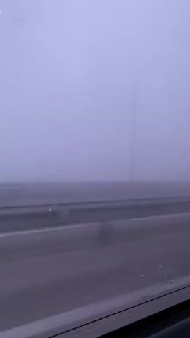 The video shows a view from a vehicle moving through dense fog. The scene is dominated by a muted purple-gray sky and fog that obscures the horizon and any distant objects. In the foreground, there is a highway with a central barrier, likely made of metal, running horizontally across the frame. The road surface appears wet, with droplets of water visible on the window through which the video is recorded. The fog is so thick that it is difficult to discern any details of the landscape beyond the immediate road and barrier. A faint vertical pole, possibly a lamppost, is visible in the distance on the left side of the frame, almost completely swallowed by the fog. The video captures the experience of traveling in low visibility conditions.