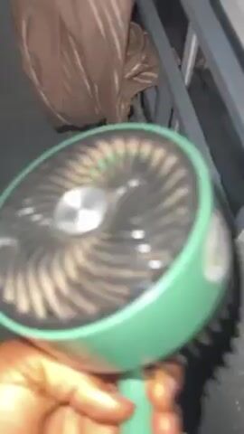 A hand holds up a dirty, hand-held fan. The fan is round with a light green outer ring and a silver blade assembly within. A white circle is located in the center of the blade assembly. The fan blades are covered in dust. The background is blurry and indistinct.