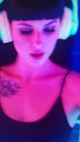 A woman with pale skin and dark hair and bangs is working as a DJ in front of a digital mixing console, with swirling, color-changing visual effects in the background. She wears a black spaghetti-strap tank top and white headphones with glowing blue-green lights on them. The woman has tattoos on her left arm and right upper chest. The background features a variety of special effects, from green flames to pixelated squares and clouds. The lighting casts a purple and pink hue on the scene. The music has a strong techno beat.