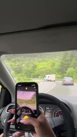 Here is a description of the video:
The view is from inside a car, looking out the windshield as it drives on a winding road lined with green trees. Ahead are two vehicles: a white van and a gray car. The driver is holding a black iPhone in their left hand, recording a video of the road ahead, mirroring the view through the windshield. The dashboard and steering wheel of the car are visible. The needle of the speedometer points to 25. The interior of the car is gray. The driver has pink manicured fingernails. The road appears slightly wet.