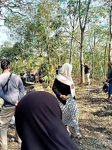 The video shows a gathering of people outdoors in a wooded area with some structures visible. In the foreground, a person wearing a black headscarf is facing away from the camera. To the right, a woman wearing a light-colored hijab, black jacket, and patterned pants is walking and looking to her left. Several other individuals are scattered throughout the scene, some sitting, some standing. In the background, there is a building with a roof, and many more people are gathered around it. The environment appears to be dry and sunny with trees and vegetation.