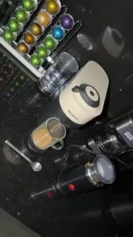 On a black, speckled countertop sits a Nespresso coffee maker, with a rack of colorful coffee pods to the side. A glass of coffee with some contents already inside sits next to a small spoon.  Nearby is a milk frother with foam inside. Two empty clear glasses and another appliance are also visible. The scene is well-lit.