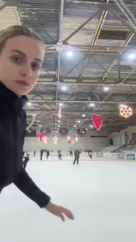 The video shows a woman ice skating indoors. She has blonde hair and is wearing a black jacket and pants, and a black belt. She moves across the ice, holding her arms out for balance. There are several other ice skaters in the background. The ice rink has hanging decorations, including hearts and snowflakes, and overhead lights. The roof of the building is exposed and appears to be industrial.