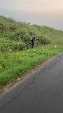 A lone figure wearing a helmet stands on the side of a paved road, surrounded by tall green grass and vegetation. The figure appears to be holding a phone and looking at it. The road curves to the right, and a blurred landscape of hills and vegetation is visible in the background. The sky is overcast.