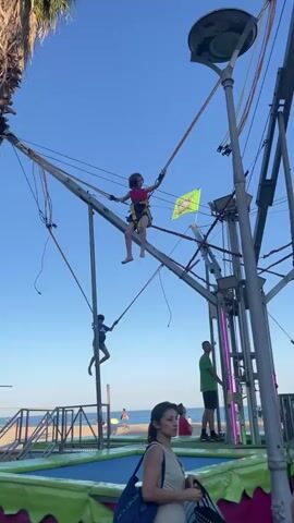 This media file captures an outdoor scene at what appears to be a beachside amusement area or fair. The main subject is a bungee trampoline attraction, where children are seen jumping high into the air, secured by harnesses and bungee cords.

In the foreground, a woman with dark hair and a light-colored top is looking to the side. She is holding a bag with a white and black polka dot pattern and another blue bag over her shoulder. Behind her, another person with dark hair and glasses is visible, wearing a black backpack.

The bungee trampoline structure is prominent, with metal poles, cables, and harnesses. A bright green flag with yellow lettering is attached to one of the poles, and it appears to read "Happy Jumping." Another similar flag is visible further back. Several children are actively using the attraction, reaching considerable heights. One child in a red and yellow harness is particularly visible in mid-air. Another child in a blue shirt is also jumping.

In the background, there is a glimpse of the sea, suggesting the location is near a beach. The sky is clear and blue, indicating pleasant weather, likely during the daytime, possibly late afternoon or early evening given the lighting. A few other people are casually observing the activity. A man in a green t-shirt and dark shorts stands near the structure, possibly an attendant or spectator. Another person is partially visible further back, and a man with a beard is seen on the right side of the frame. Pink lights are integrated into the structure, adding a festive touch. The overall atmosphere seems lively and enjoyable, with children experiencing the thrill of jumping.