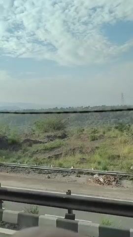 The video shows a view from a vehicle, likely traveling on a highway. The landscape is hilly and covered in sparse green vegetation. There are signs of human construction, including what appears to be a railway track and a road barrier. In the distance, there are power lines and a transmission tower. The sky is blue with scattered clouds. The video is shot from a moving perspective, indicating the viewer is observing the scenery as they pass by. There are no people visible in the frame.