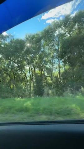 The video is shot from the interior of a moving vehicle. The view through the side window shows trees and green grass. The sun is visible through the trees, casting bright rays. The car appears to be on a road with a clear blue sky and white clouds above. The interior of the car, including the dashboard and a side mirror, is also visible. There are no people visible in the video.