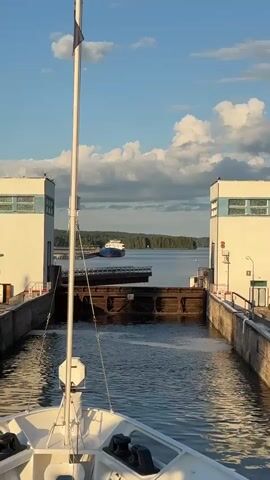 The video shows a body of water with a large ship in the distance. In the foreground, the bow of a white boat is visible, with a flagpole and a white antenna. The boat appears to be approaching a lock, with two large white structures on either side and a brown gate in the water. The sky is blue with white clouds. The scene is well-lit, suggesting it is daytime.