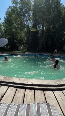 A group of people are swimming and splashing in a round pool on a sunny day. The pool is surrounded by a wooden deck, and trees and foliage are visible in the background. Some people are standing in the water, while others are swimming. The water is a clear turquoise color and is disturbed by the movements of the swimmers. The atmosphere appears to be relaxed and joyful, with people enjoying the warm weather and the opportunity to cool off.