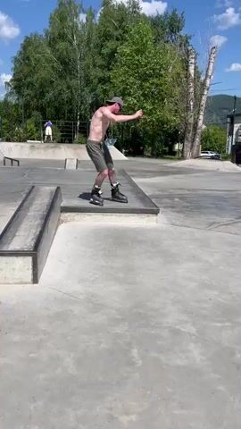 The video shows a person on inline skates in a skatepark on a sunny day. The person is wearing a hat, sunglasses, and shorts, and is shirtless. They are in the process of performing a trick, with their body angled and arms extended. The skatepark appears to be made of concrete, with ramps and other obstacles visible. Trees and a hillside are in the background, suggesting an outdoor, possibly mountainous location.