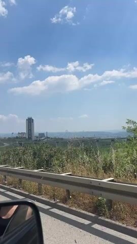 The video shows a long-distance view of a large, white cable-stayed bridge spanning a considerable distance. The bridge is framed by a foreground of greenery and a lower-level highway bridge. The sky is a light blue with scattered white clouds. In the background, hazy mountains are visible.  The perspective suggests the video was taken from a moving vehicle, perhaps another highway, given the guardrail visible in the lower frame.