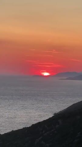 The video shows a sunset over the ocean. The sun is a bright red orb on the horizon, casting a warm glow across the sky and water. The sky is a gradient of orange and pink, with some wispy clouds. The ocean is a deep blue, with gentle waves. In the foreground, there is a green, hilly landscape. The camera is positioned high up, looking down on the scene.
